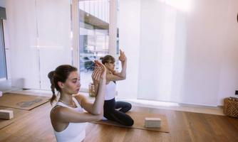 Two women are practicing yoga in a bright room. They are on mats and focusing on their breathing.