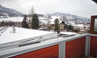 A view from a balcony of a snowy landscape with mountains in the background. The surroundings are calm and wintry, with houses and trees in the foreground.