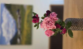 A bouquet of pink and dark red flowers stands in a decorative vase. In the background, there is a landscape with mountains and a meadow.