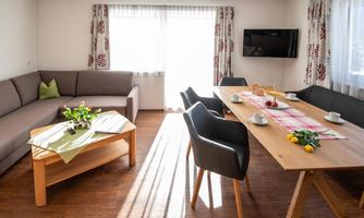 A bright living room with a cozy sofa and a dining table. On the table, there are flowers and dishes, and large windows let in a lot of light.