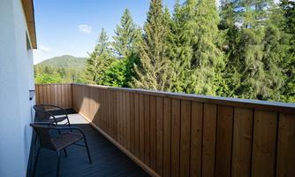 A balcony with a wooden railing and two chairs. In the background, green trees and gentle hills can be seen.