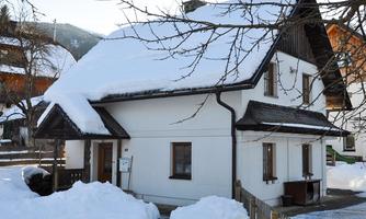 A cozy house in the snow with a snow-covered roof. The surroundings are quiet and wintry, with snow-covered trees and a clear sky.