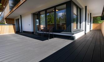 A modern balcony with a bright wooden railing and two chairs. The floor is dark and the large front window offers a view of the greenery.