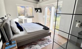 A modern bedroom with light walls and wooden ceilings. There is a large bed, a nightstand, and large windows that let in plenty of light.