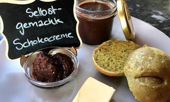 A plate with homemade chocolate cream, a jar of chocolate sauce, and two small rolls. Also included are a piece of butter and some cookies.