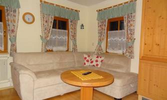 A cozy living room with a beige couch and a round table. The windows are adorned with curtains, and a wooden cabinet stands in the corner.