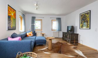 A modern living room with a blue couch and colorful cushions. The room is bright and features large windows as well as stylish artworks on the walls.