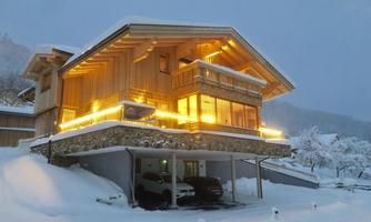 A modern chalet in the snow, illuminated by warm lights. The surroundings are wintry with plenty of snow and a foggy sky.