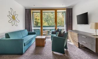 A modern living room with a blue sofa, a wooden table, and two armchairs. Large windows let in plenty of light and offer a view of the greenery.