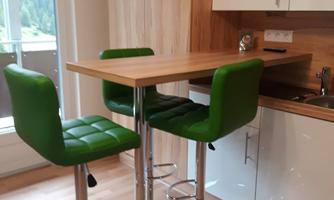 A modernly furnished kitchen with a small wooden bar table and three green bar stools. The atmosphere is bright and inviting.