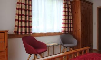A cozy room with a window through which daylight streams. Two chairs, red and grey, stand next to a small table.