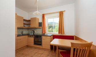A modern kitchen with wooden cabinets and a dining area. The windows offer a view of nature.
