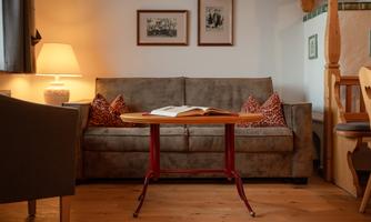 A cozy living room with a gray sofa and two decorative cushions. On a table lies an open book, next to it stands a lamp, and the walls are adorned with pictures.