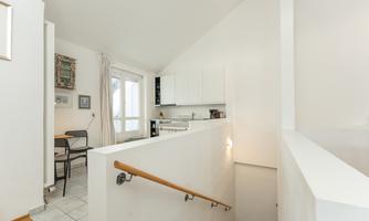 A bright and modern kitchen with white furniture and large windows. On the left is a small dining area with a table and chairs.