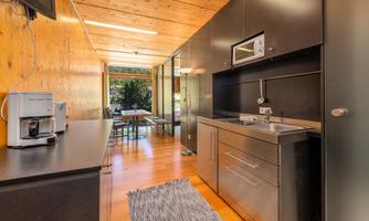A modern kitchen with wood and metal elements. In the background, there is a table with chairs and a large glass front that opens up the view to the outside.