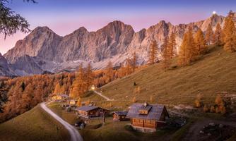 A picturesque mountain landscape with vibrant orange trees in autumn. In the foreground, traditional wooden chalets and a winding path can be seen.