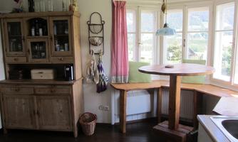 A cozy kitchen with a wooden shelf and a round dining area. Large windows offer a view of the landscape.