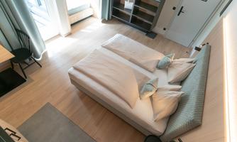 A modern bedroom with a large bed and light wooden floor. The windows let in plenty of daylight and the room is furnished in a minimalist style.