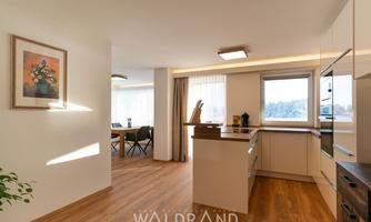 Modern, bright kitchen with wooden floor and large windows. A dining area is visible in the background.