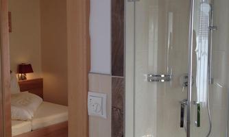 A modern bathroom with a glass shower and bright tiles. In the background, a cozy bedroom with a double bed can be seen.