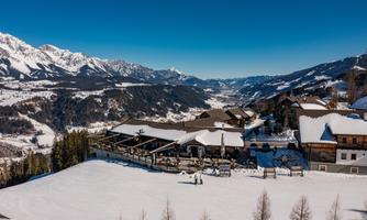 A snowy mountain landscape with a cozy cabin and guests on the terrace. In the background, majestic mountains and a clear blue sky can be seen.