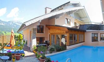 A modern house with a beautiful swimming pool and flower pots. In the background, the mountains and a blue sky can be seen.