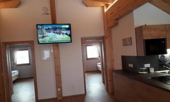 A cozy interior with wooden details and a modern kitchen corner. A television is hanging on the wall, and there are two bedrooms visible with windows.