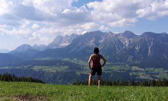 A person stands in a meadow, looking at impressive mountains. The sky is partly cloudy, and the landscape appears very picturesque.