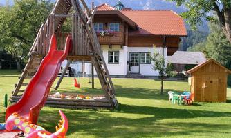 A beautiful garden with a playground that includes a slide and a playhouse. In the background, there is a traditional house and the mountains are visible.