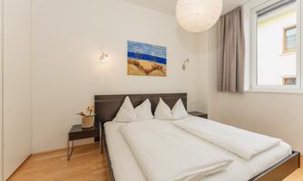 A cozy bedroom with a large bed and light wooden flooring. A picture of a beach landscape hangs on the wall, and the window lets in natural light.