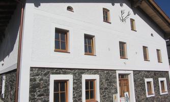 A traditional building with white walls and brown window frames. The ground floor is made of stone and features a wooden porch.