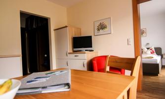 A cozy living space with a dining table and chairs. In the background, a television and a sofa can be seen.