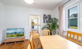 A bright dining room with a wooden table and chairs. In the background, there is a television and a plant that makes the room look inviting.