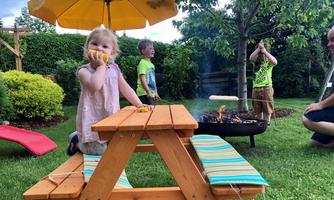 A childlike picnic in the garden, with a little girl at the table and other children nearby. A fire is burning on the grill while the sun is shining.