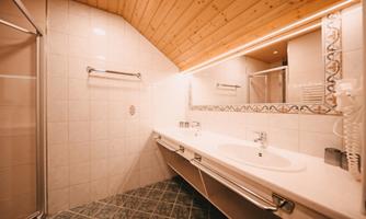 A modern bathroom with a wooden ceiling and white tiles. It features a large mirror, a double sink, and a separate shower cubicle.