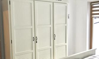 An elegant, white wardrobe with three doors stands in a modernly furnished bedroom. In the foreground, there is a bed with checkered bedding.