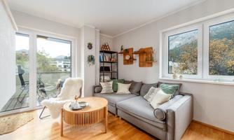 A cozy living room with a gray sofa and a round wooden table. Large windows provide a view of nature and ensure plenty of daylight.