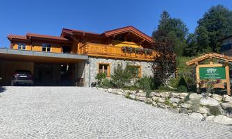 A modern wooden house with a rock garden and a paved driveway. The sky is clear and blue, surrounded by trees.
