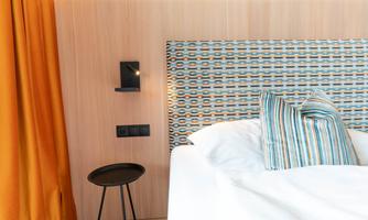 A modern bedroom with a comfortable bed and a colorful headboard. The orange curtains and the minimalist furniture create a cozy atmosphere.