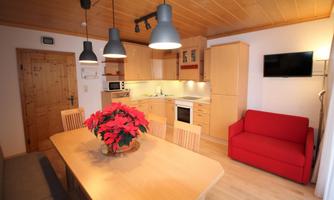 A modern kitchen corner with a large dining table and a red sofa. The walls are light, and the furniture is made of light wood.