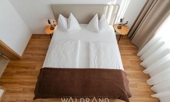 A modern bedroom with a double bed and a striking bedspread. The room has a warm wooden floor and large windows that let in daylight.