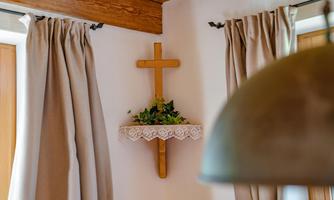 A wooden square with a carved cross and green plants on it. The background features bright curtains and a warm wooden ceiling.