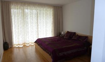 A modern bedroom with a large bed and purple bedding. Bright curtains let in a lot of light.