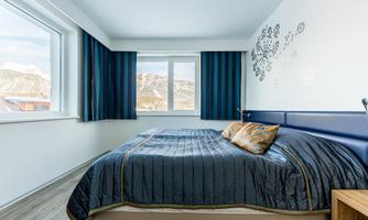 A modern bedroom with a large bed and blue curtains. A mountain landscape can be seen through the windows.