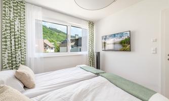 A modern bedroom with a double bed and bright curtains. The green landscape is visible through the window, and a television is hanging on the wall.