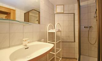 A modern bathroom with tiled walls and a sink. On the left is a shower with a transparent shower partition and a shelf for towels.