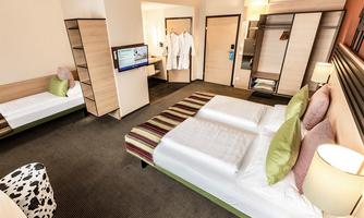 A modern hotel room with two beds and a single bed. The decor is bright and inviting, with a television and comfortable seating.