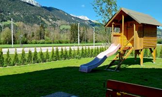 A playhouse with a slide stands on green meadows, surrounded by mountains and a blue sky. On the table, there is a jug and a flower vase.