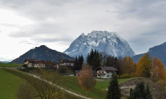 A picturesque mountain landscape with snow-capped peaks in the background. In the foreground, houses and trees in autumn colors can be seen.