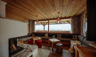 A cozy living room with a wooden ceiling and large windows. There is a sitting area with a couch and a fireplace, as well as bright lamps and a rustic table.